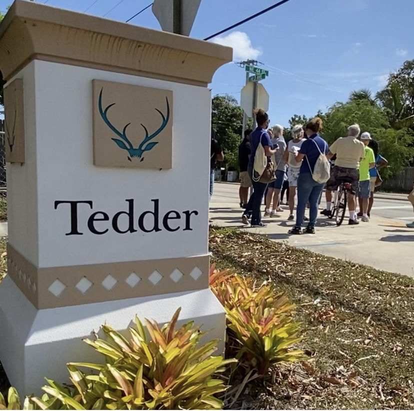 Attendees stand on a sidewalk in the background, a Tedder community sign sits in the foreground.